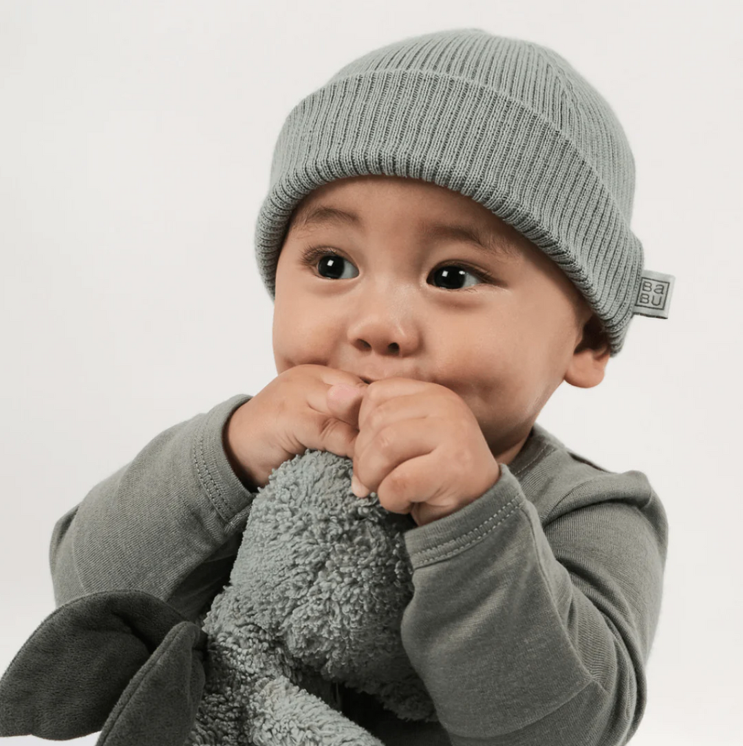 A baby wearing a gray knit beanie and gray outfit holds a plush toy, gently biting on its hands. The background is plain white.