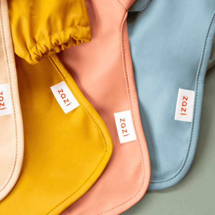 Three Zazi Recycled Full-Sleeved Bibs, each in yellow, peach, and blue with the "Zazi" label, are arranged in a fan-like display on a soft, neutral background.