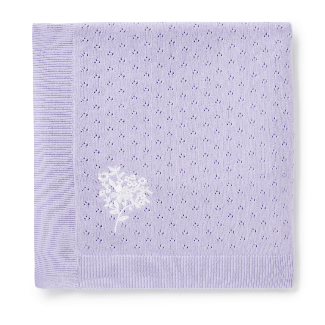 A folded, lavender-colored Aster & Oak Organic Ruffle Knit Baby Blanket made from organic cotton by Aster & Oak, featuring an embroidered white floral design on one corner and a ribbed edge.