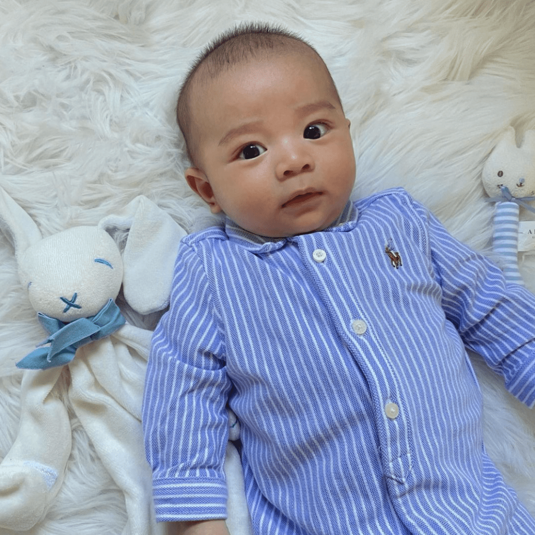 A baby in a blue striped onesie lies on a soft white surface, flanked by two stuffed toys made from organic cotton: the Maud N Lil Organic Bunny Comforter - Gift Boxed on the left and a white cat on the right. This cozy scene makes an adorable baby gift.