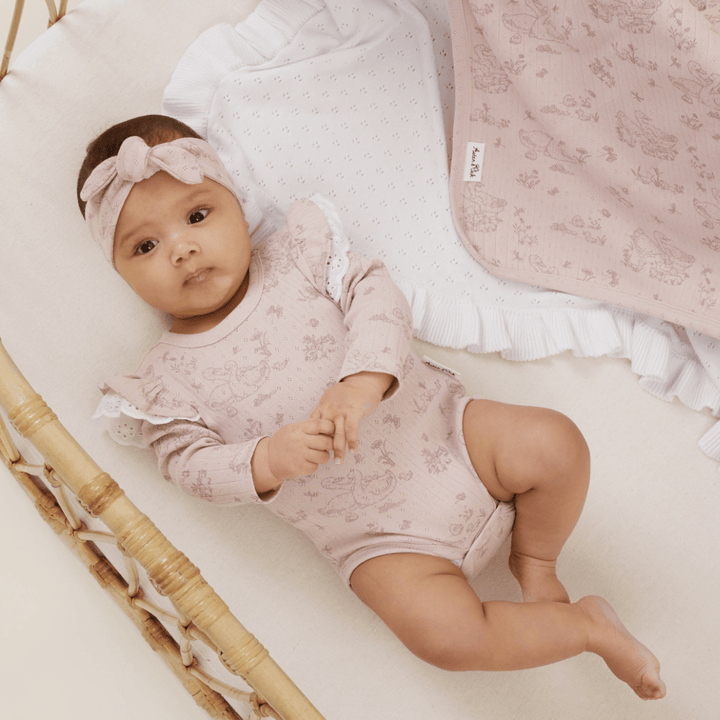 A baby wearing a pink outfit and headband lies in a wicker bassinet. Next to the baby is an Aster & Oak Organic Ruffle Knit Baby Blanket.