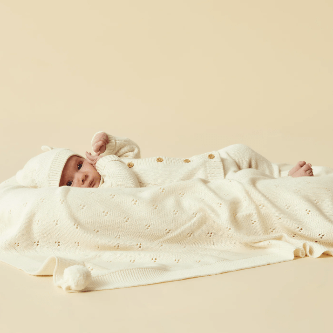 An infant dressed in a white outfit and hat lies on a mint-green Wilson & Frenchy Knitted Pointelle Baby Blanket, offering an adorable scene perfect for a baby shower gift. The beige background accentuates the heirloom design, adding a timeless touch.