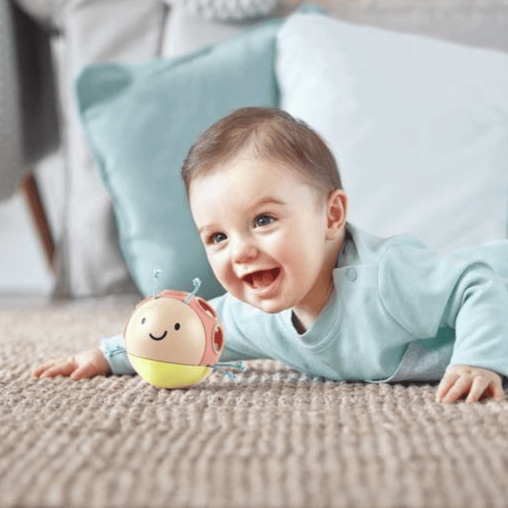 A baby in a light blue outfit lies on a carpet next to a small, smiling, ladybug-shaped Hape Busy Bug Roll by Hape, encouraging tactile development.