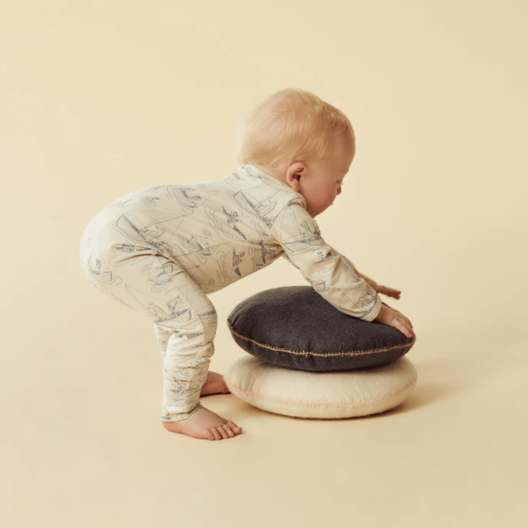 A newborn in Wilson & Frenchy organic baby pyjamas adorned with hand-illustrated prints crouches on stacked cushions against a beige background.