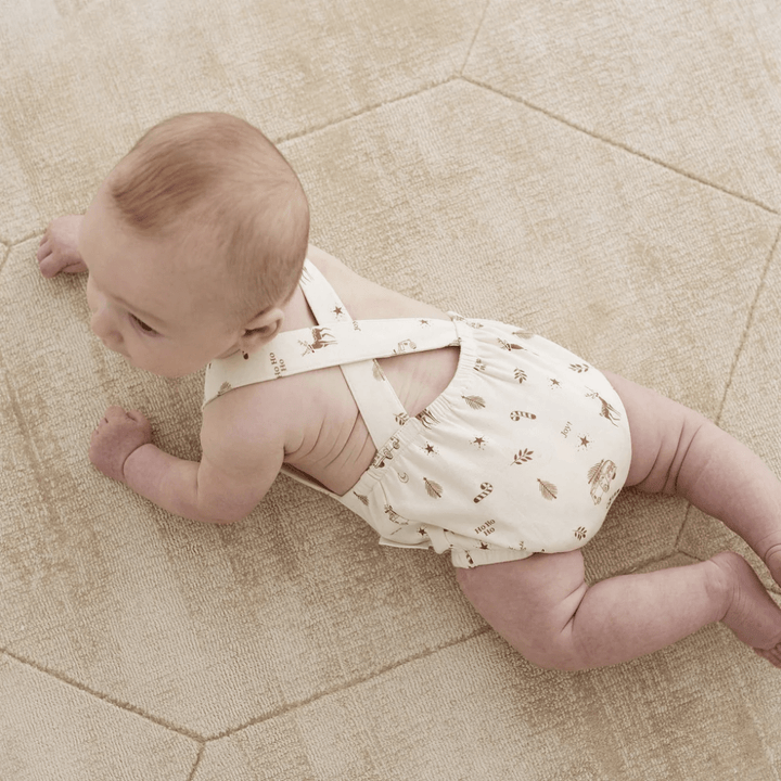 A toddler clad in a snug Aster & Oak Organic Happy Holidays Playsuit - LUCKY LAST - 2 YEARS ONLY explores on a textured beige carpet, inquisitively gazing to the side.