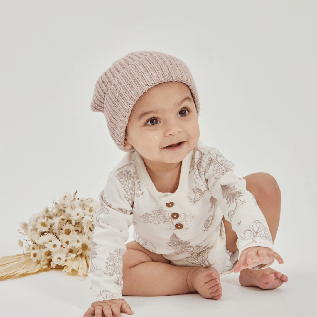 A baby wearing a cozy knit hat and an Aster & Oak Organic Henley Long-Sleeved Onesie in the FOX WOODS design sits on the floor next to a bouquet of dried flowers.