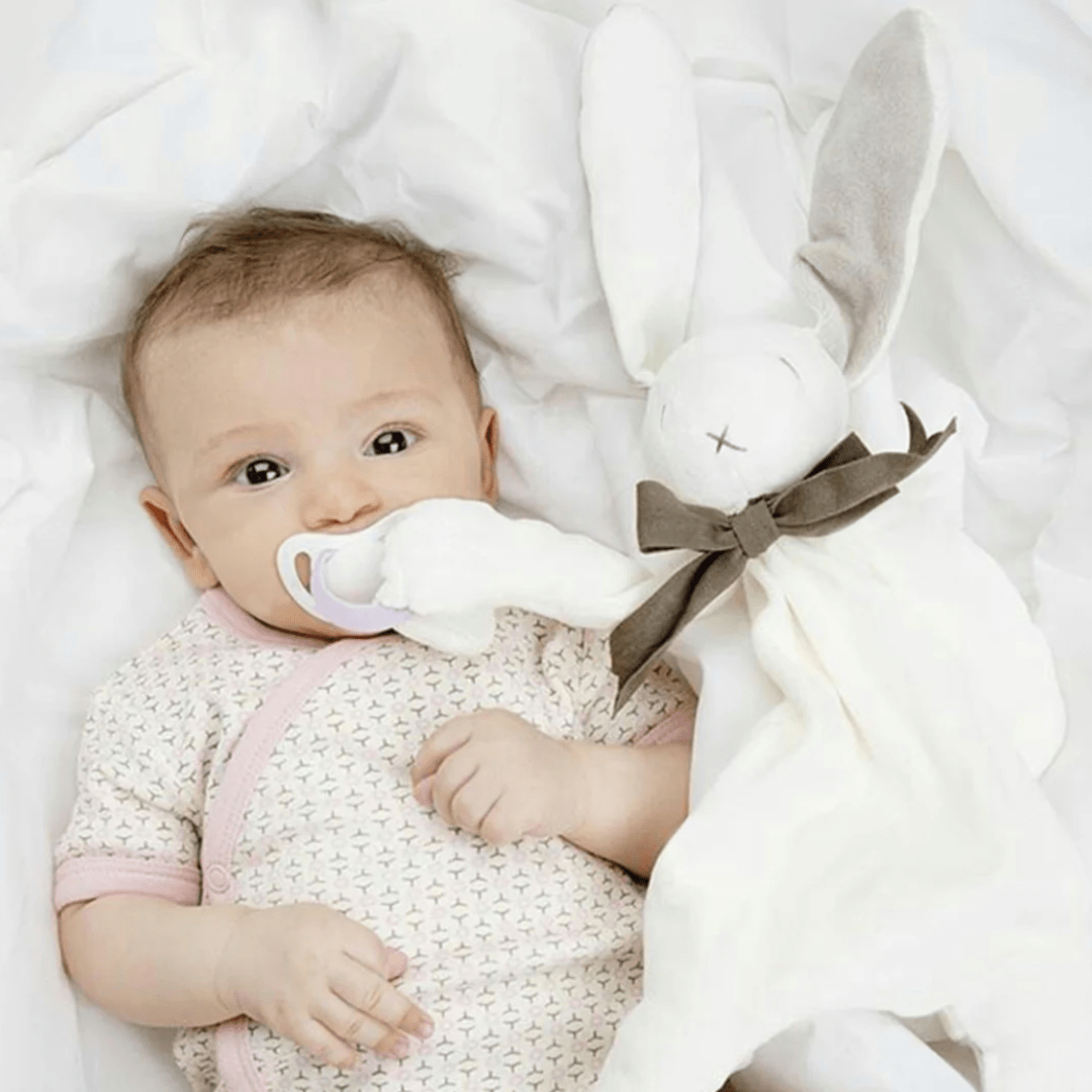 A baby lying on a white blanket holds a pacifier and the Maud N Lil Organic Bunny Comforter – Gift Boxed, making it an ideal baby gift.