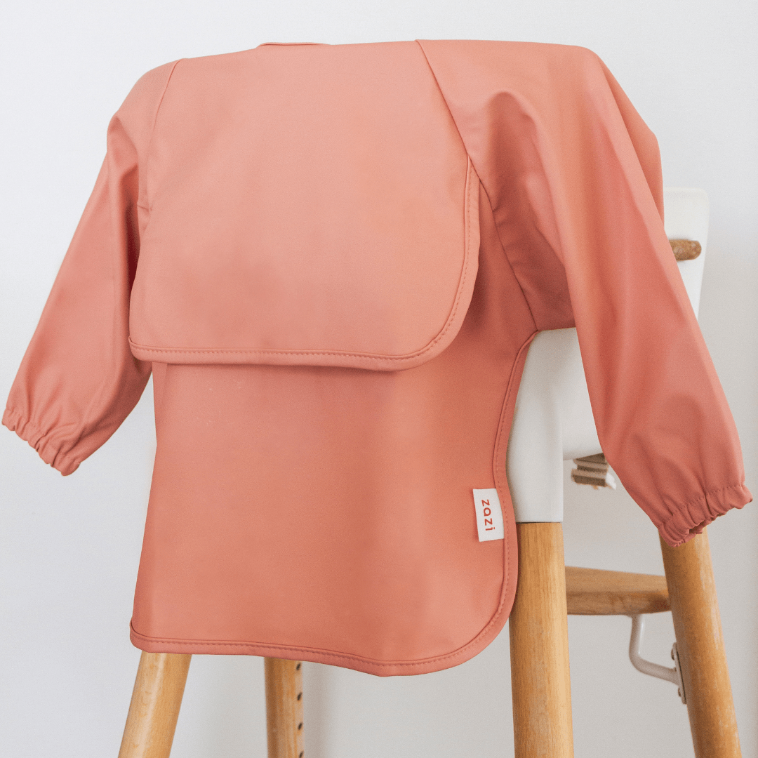 A Zazi Recycled Full-Sleeved Bib in pink, which is machine washable, hangs on a wooden high chair. It has a label reading "2021" on the side.