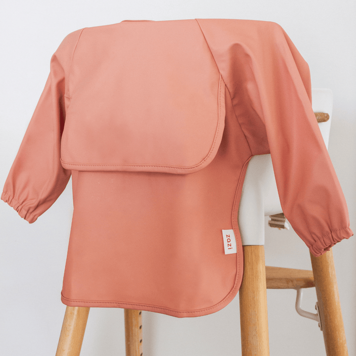 A Zazi Recycled Full-Sleeved Bib in pink, which is machine washable, hangs on a wooden high chair. It has a label reading "2021" on the side.