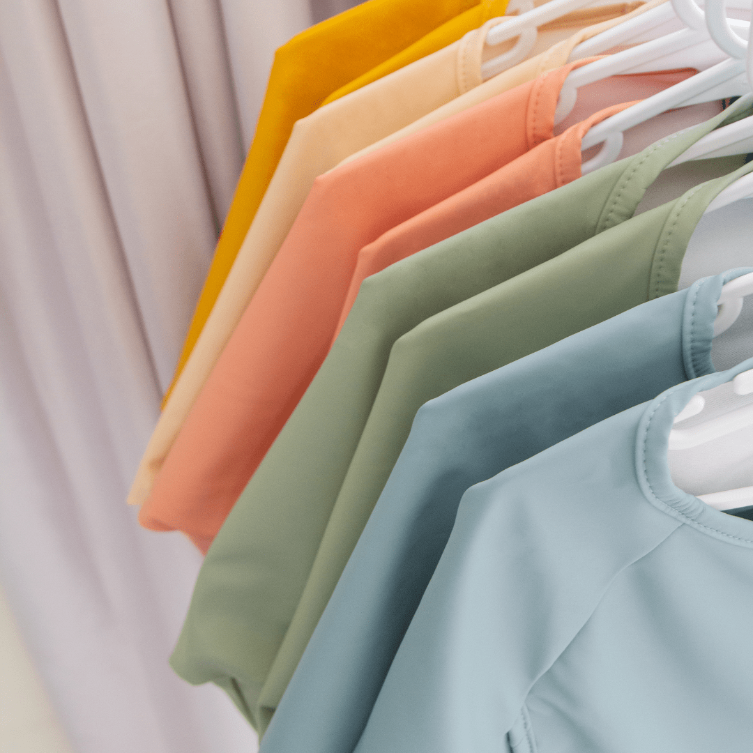 A row of colorful, neatly hung shirts on white hangers in shades of yellow, orange, peach, green, and blue. Adding a playful touch to the scene are Zazi Recycled Full-Sleeved Bibs by Zazi, which are machine washable.