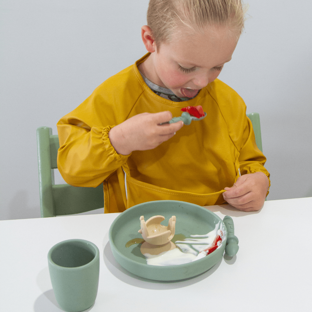 A child in a yellow Zazi Recycled Full-Sleeved Bib from Zazi eats food messily from a green plate with a green cup beside it.