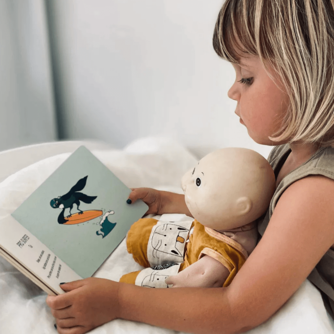 A little girl reading a "Kiwi Surfers" Board Book by As We Are Illustration to a doll in a local spot.