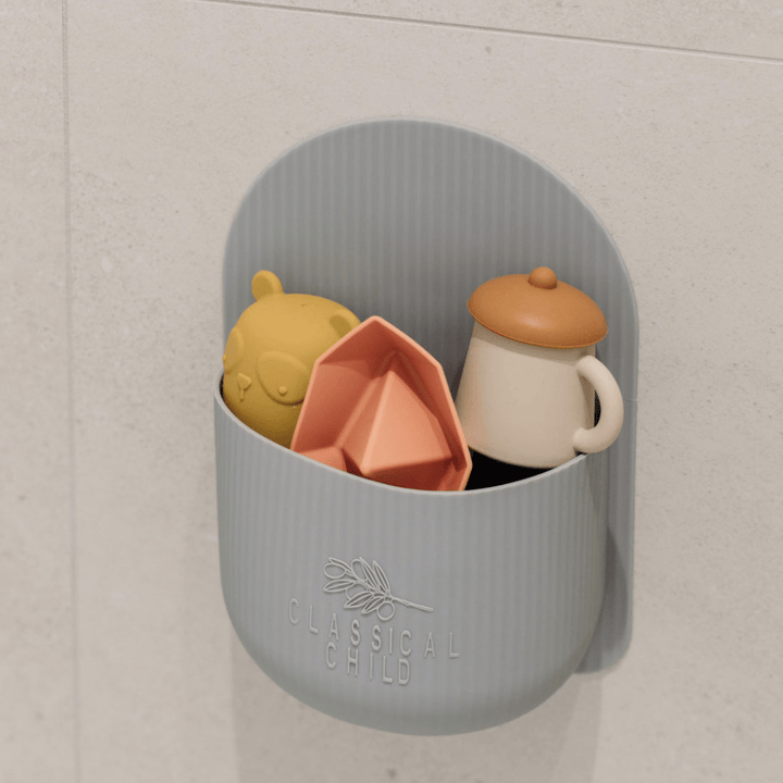 Wall-mounted gray holder crafted from BPA-free silicone, featuring toys like a yellow bear head, an orange boat, and a white pitcher with a brown lid—ideal for organizing bath toys as part of the Classical Child Silicone Bath Toy Caddy - LUCKY LASTS by Classical Child.