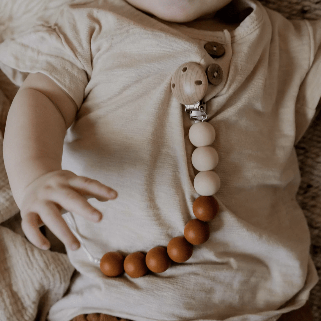 Close-up of a baby wearing a beige shirt, featuring the Funny Bunny Kids Silicone & Beechwood Dummy Clip with soothing silicone and beechwood beads in shades of brown and beige.