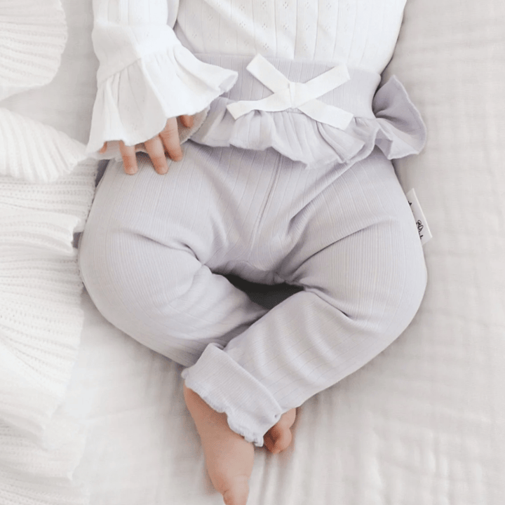 A baby wearing light-colored ruffled pants and a matching long-sleeve top, lying on a white textured blanket. The outfit includes soft and comfortable Aster & Oak Organic Rib Lavender Leggings, perfect for snuggling.