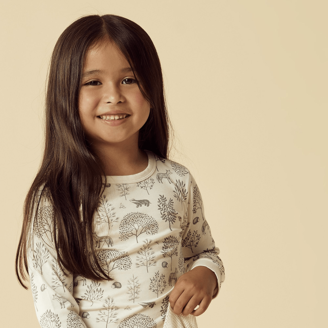 A young girl with long dark hair smiles while wearing a light-colored shirt with a nature-themed pattern against a beige background, her Wilson & Frenchy Organic Long Sleeved Pyjamas made from organic cotton adding to the adorable scene.