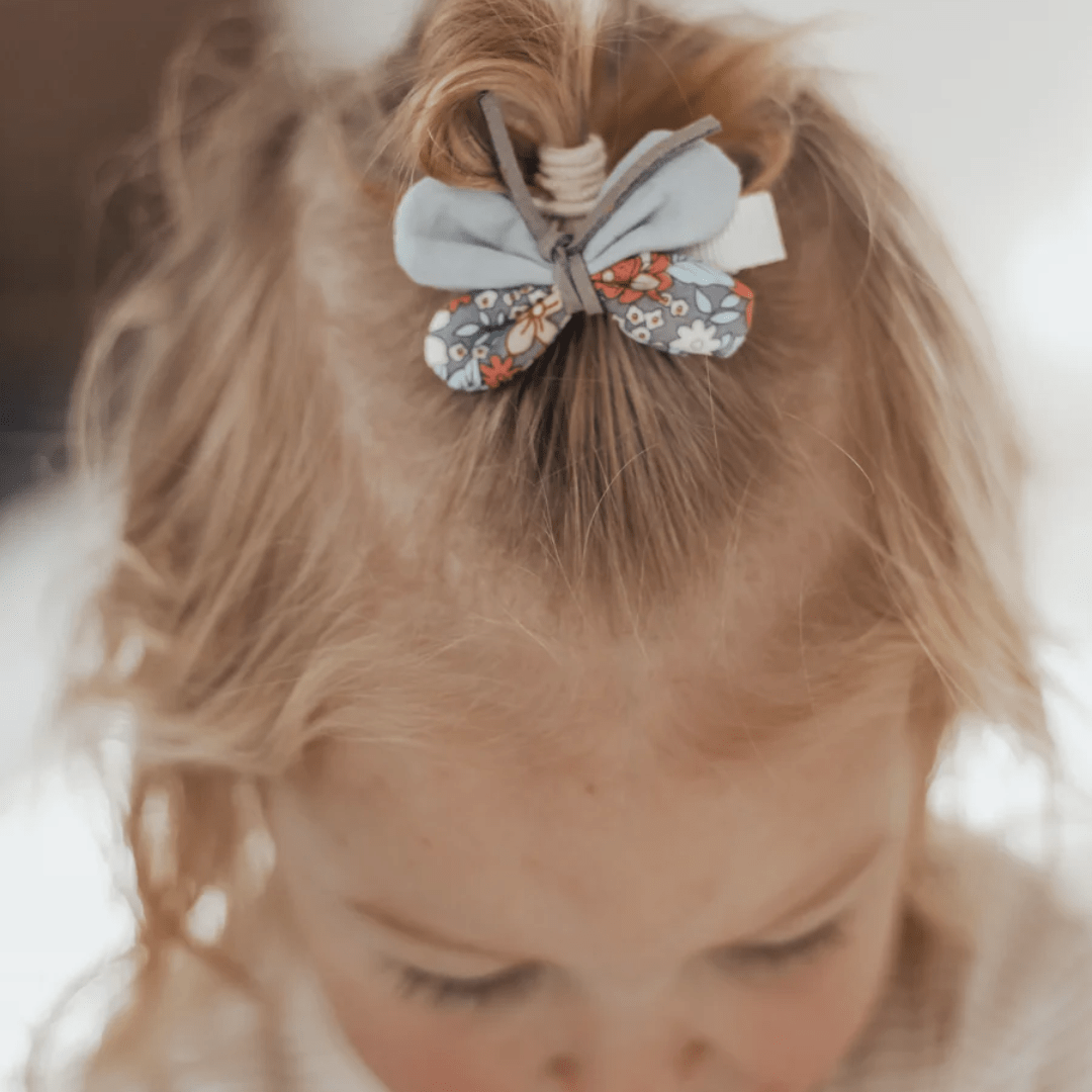 A close-up view of a child with blonde hair tied up in a ponytail, secured with Over the Dandelions Butterfly Hair Clips featuring a delicate butterfly design. The child’s face is partially visible, looking downwards.