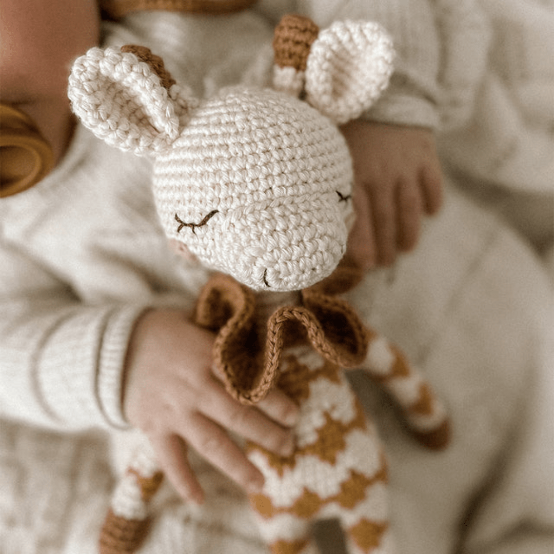 A baby holding the Patti Oslo Organic Cotton Goldie Giraffe, an eco-friendly toy crafted from organic cotton, featuring a soft textured pattern in white and brown colors.