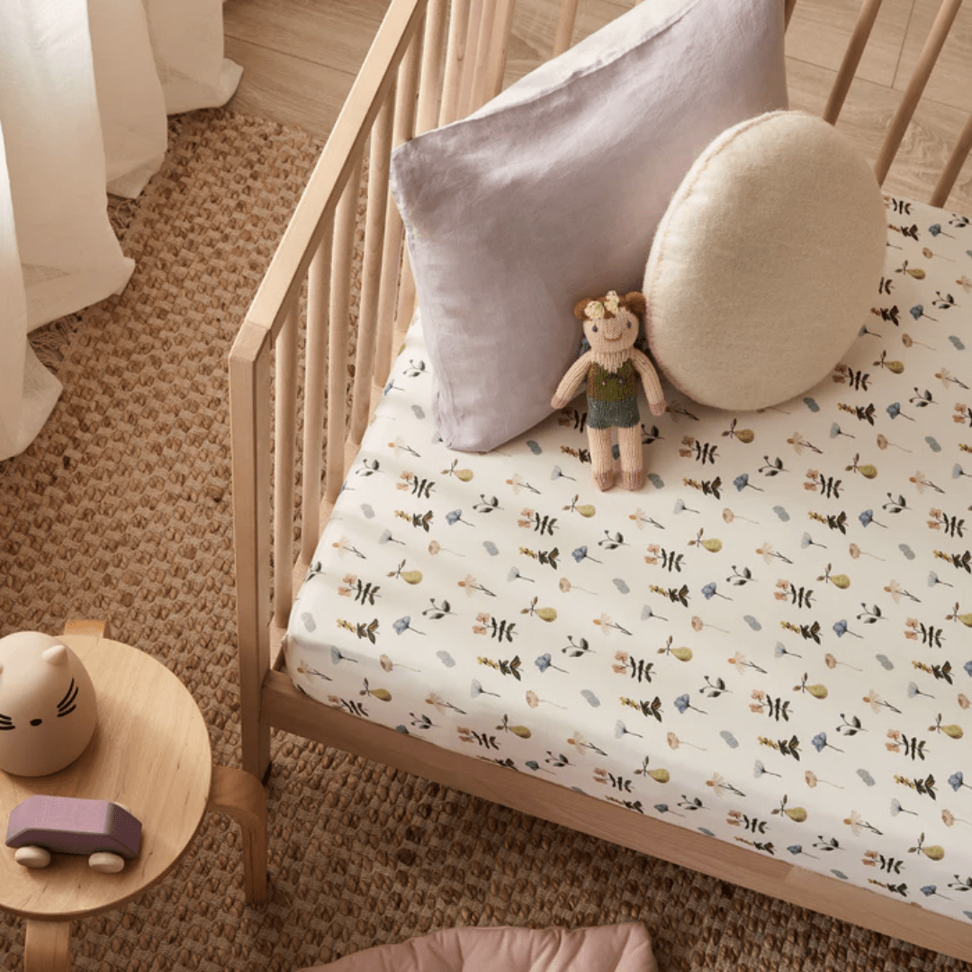 A wooden crib with a bee-themed GOTS-certified organic cotton cot sheet from Wilson & Frenchy, accompanied by a round pillow and a doll in green overalls resting against another pillow. A small wooden table nearby holds a toy cat and a purple toy car. Perfect as a baby shower gift!