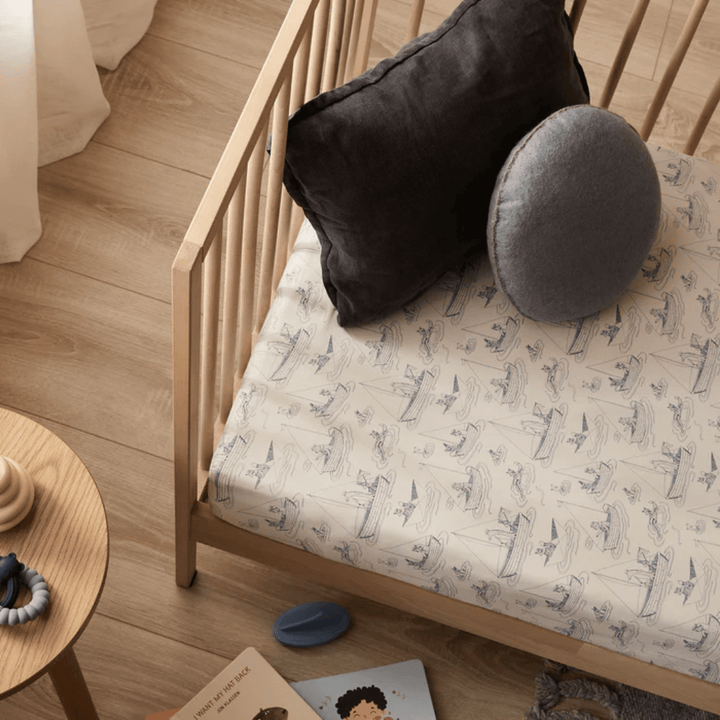 A wooden crib with dinosaur-patterned bedding, a gray round cushion, and a black rectangular pillow. A small wooden table nearby holds toys and a children's book. This setup makes for an excellent baby shower gift, especially with the addition of the Wilson & Frenchy Organic Cotton Cot Sheet in Tribal Woods.