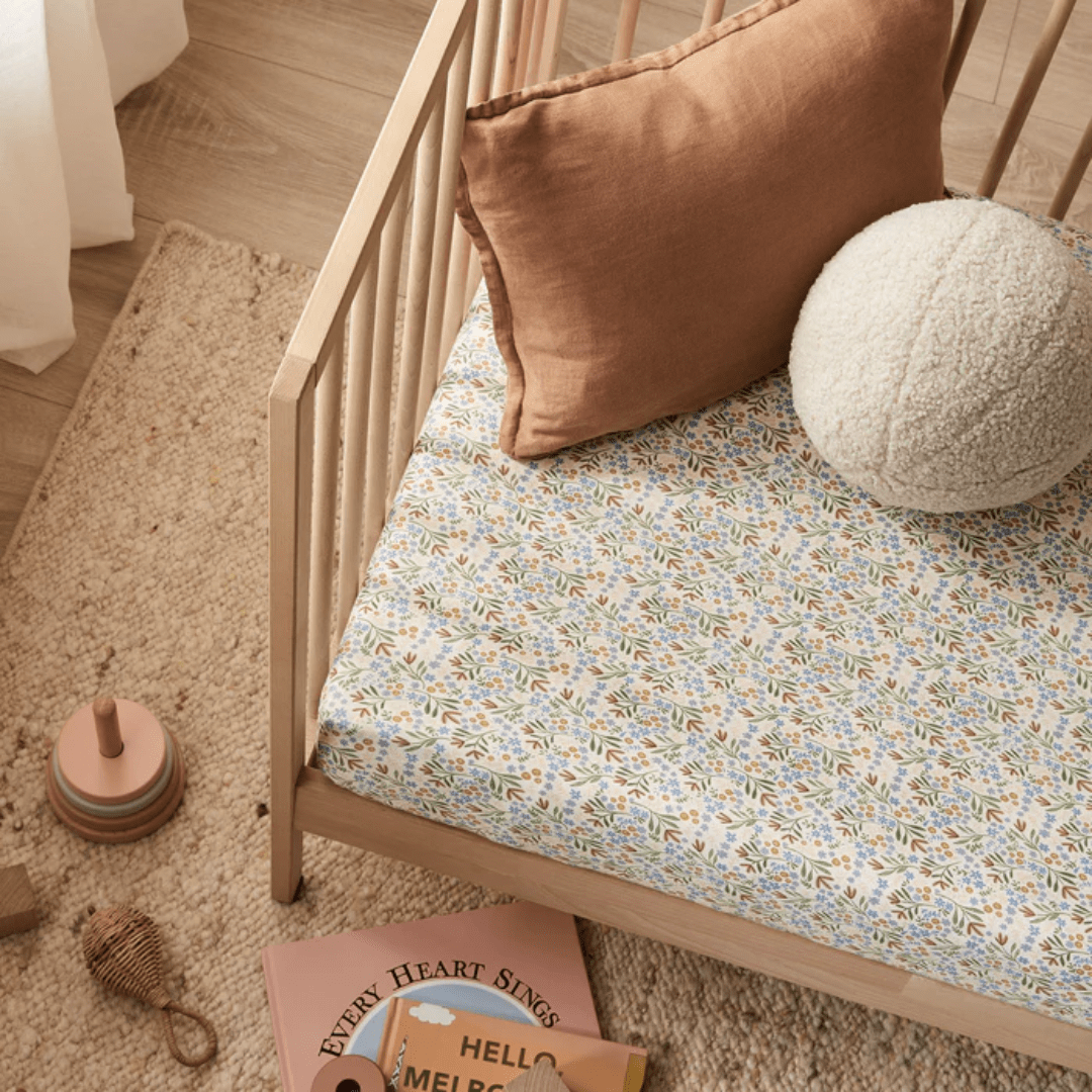 A wooden crib with a GOTS-certified Wilson & Frenchy Organic Cotton Cot Sheet in the exclusive Tribal Woods pattern and a floral-patterned pillow is shown. Nearby, there is a rug, a children's book, and some wooden stacking toys – making it the perfect setup for any baby shower gift.