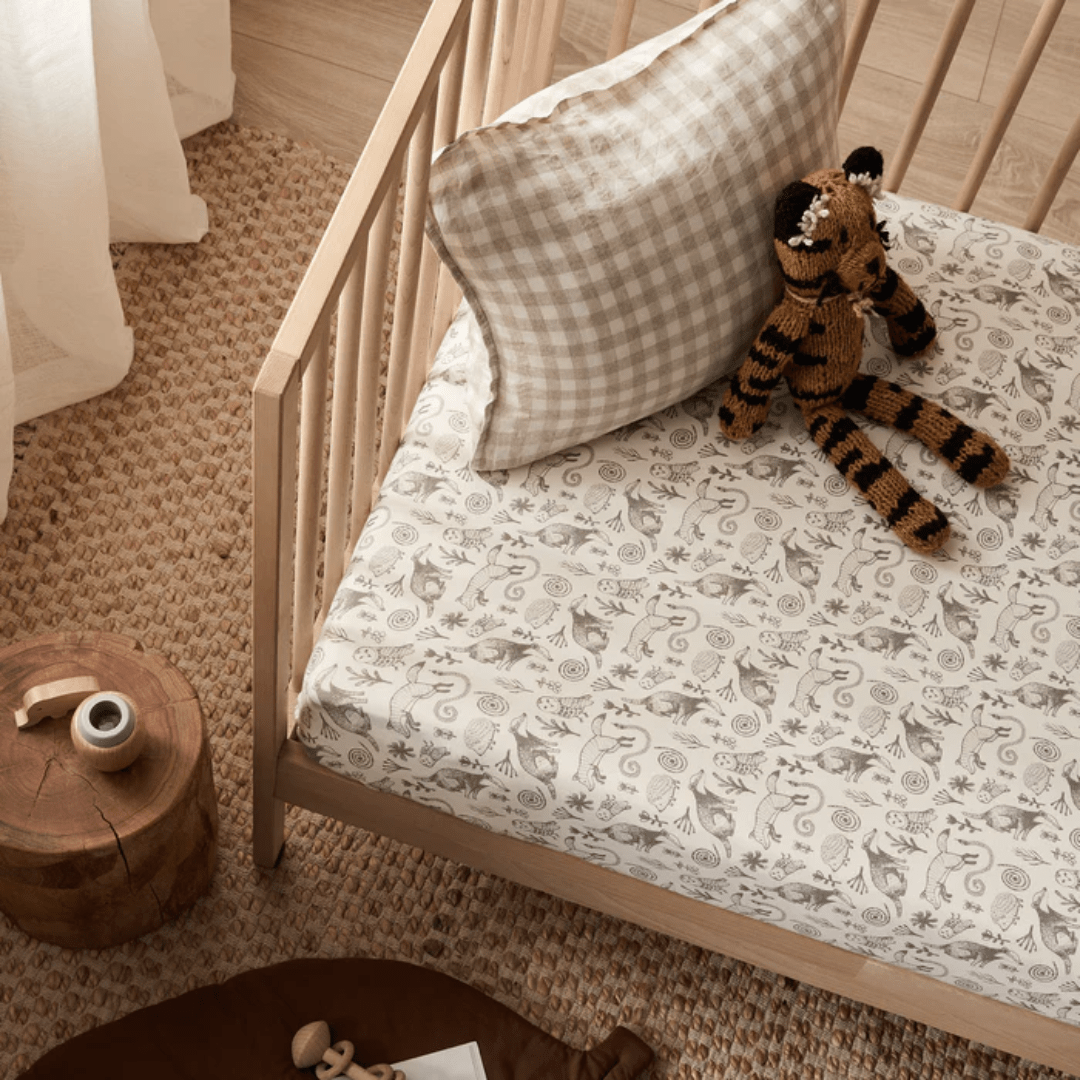 A crib featuring a checkered pillow and a toy tiger resting on the Wilson & Frenchy Organic Cotton Cot Sheet - LUCKY LAST - TRIBAL WOODS ONLY. A wooden side table with a small object and part of a mat are also visible in the room, making this cozy setup an ideal Baby Shower Gift.