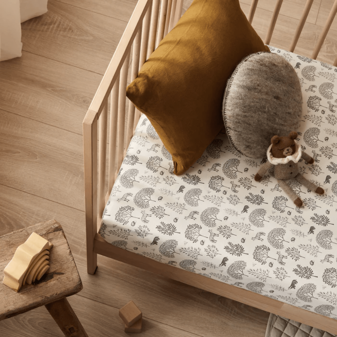 A wooden crib with patterned bedding, featuring a mustard pillow, gray cushion, and a plush bear toy. The Wilson & Frenchy Organic Cotton Cot Sheet - LUCKY LAST - TRIBAL WOODS ONLY adds a touch of comfort. A small stool next to the crib holds a wooden puzzle. Wooden floor and scattered blocks are visible—perfect as a baby shower gift!