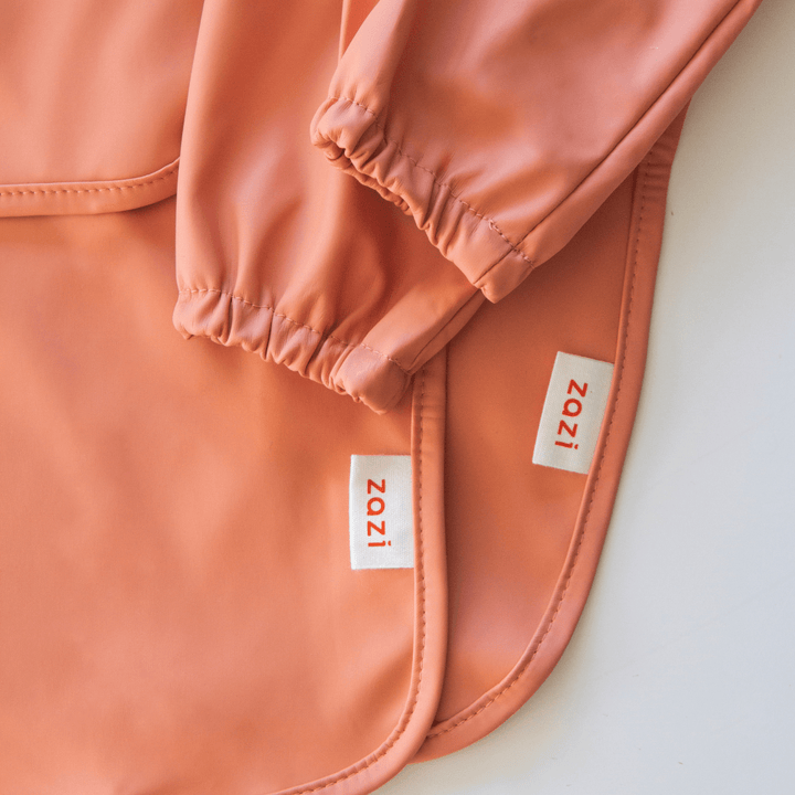 Close-up of a coral-colored, Zazi Recycled Full-Sleeved Bib featuring elastic cuffs and two beige labels displaying the text “Zazi” in red.