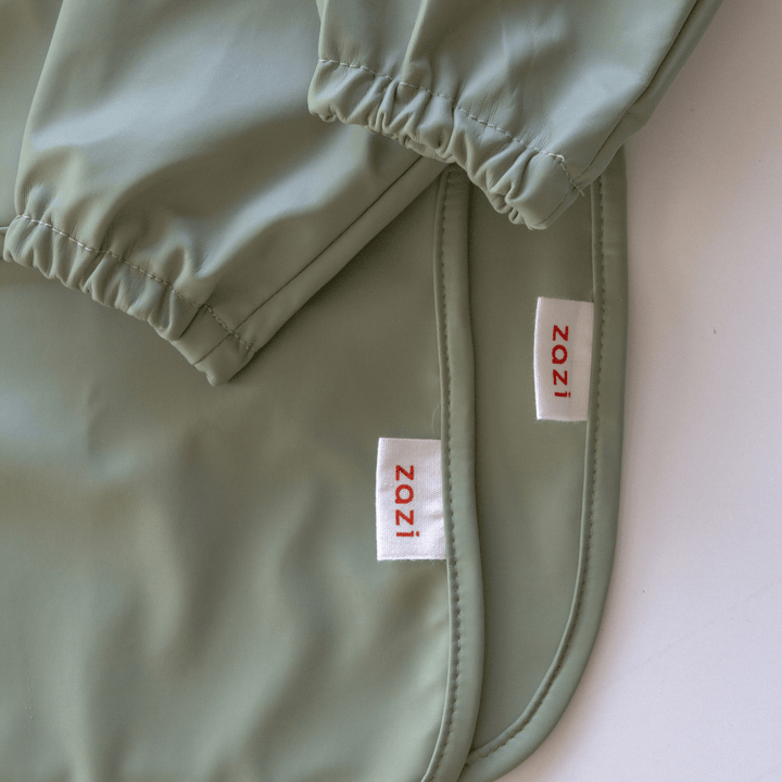 Close-up of a sage green Zazi Recycled Full-Sleeved Bib with elasticated sleeves and two white labels featuring red "Zazi" text.