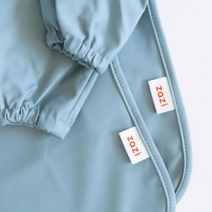 A close-up of blue-grey fabric featuring two white tags labeled "Zazi" in red text. The fabric appears to be part of a Zazi Recycled Full-Sleeved Bib.