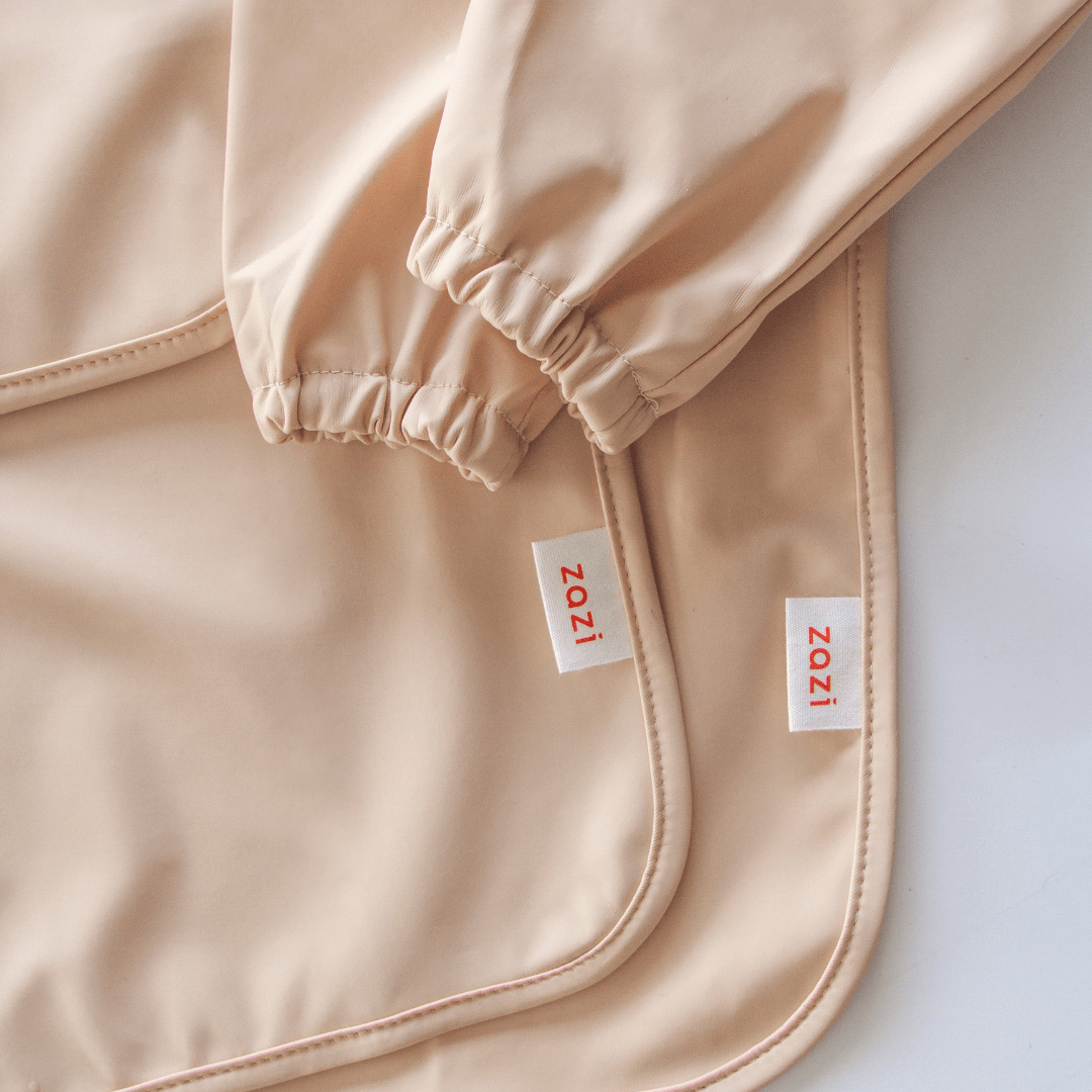 A close-up of beige fabric with elasticated cuffs and a branded tag reading "Zazi" in red text on a white background, showcasing the Zazi Recycled Full-Sleeved Bib.