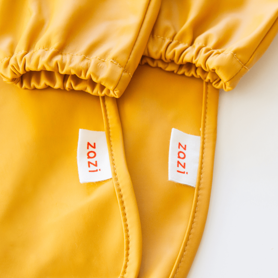 Close-up of two yellow Zazi Recycled Full-Sleeved Bibs with elastic cuffs, each labeled with a white tag reading "Zazi" in red text.