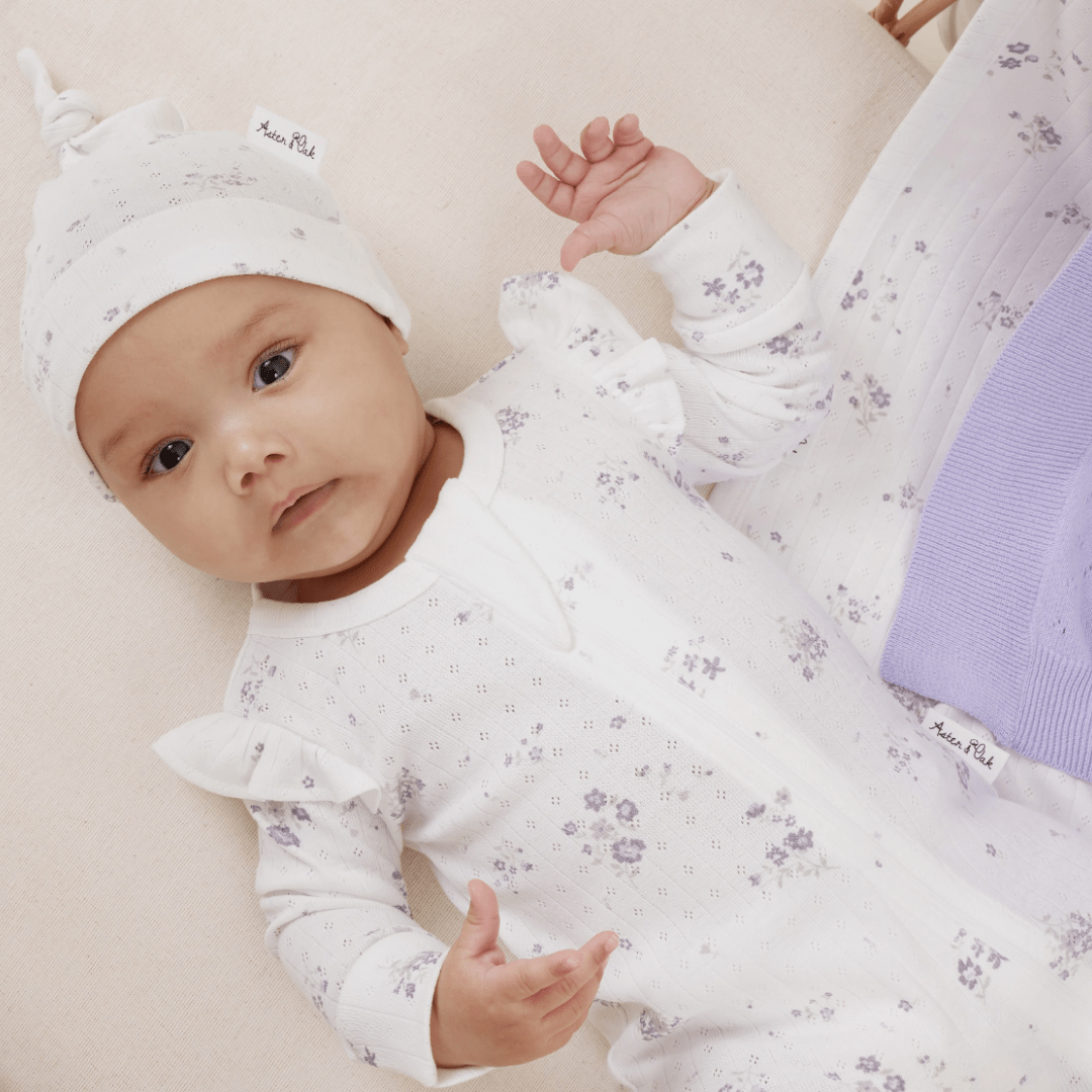 An infant lies on a beige surface wearing a white outfit with small floral patterns and matching hat, partially covered by an Aster & Oak Organic Ruffle Knit Baby Blanket from Aster & Oak.