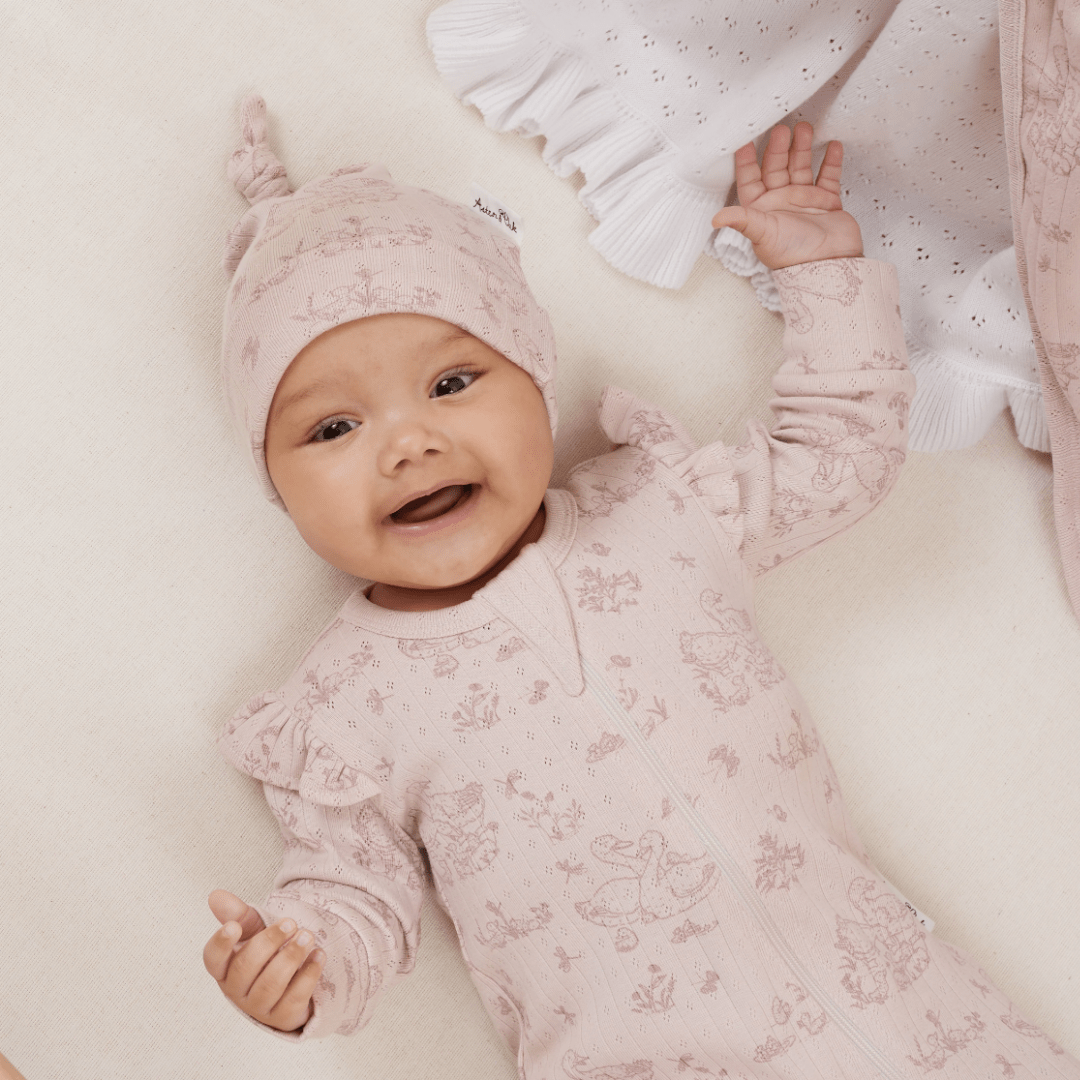 A baby wearing a pink patterned outfit and hat smiles while lying on a light-colored surface, wrapped snugly in an Aster & Oak Organic Ruffle Knit Baby Blanket by Aster & Oak.