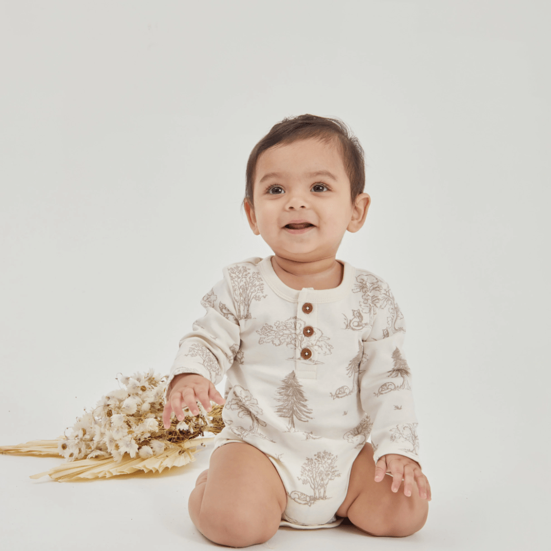 A baby sits on a white background, wearing the Aster & Oak Organic Henley Long-Sleeved Onesie in the Fox Woods design. This delightful piece of baby clothing is crafted from organic cotton and is accompanied by a small bouquet of flowers beside them.