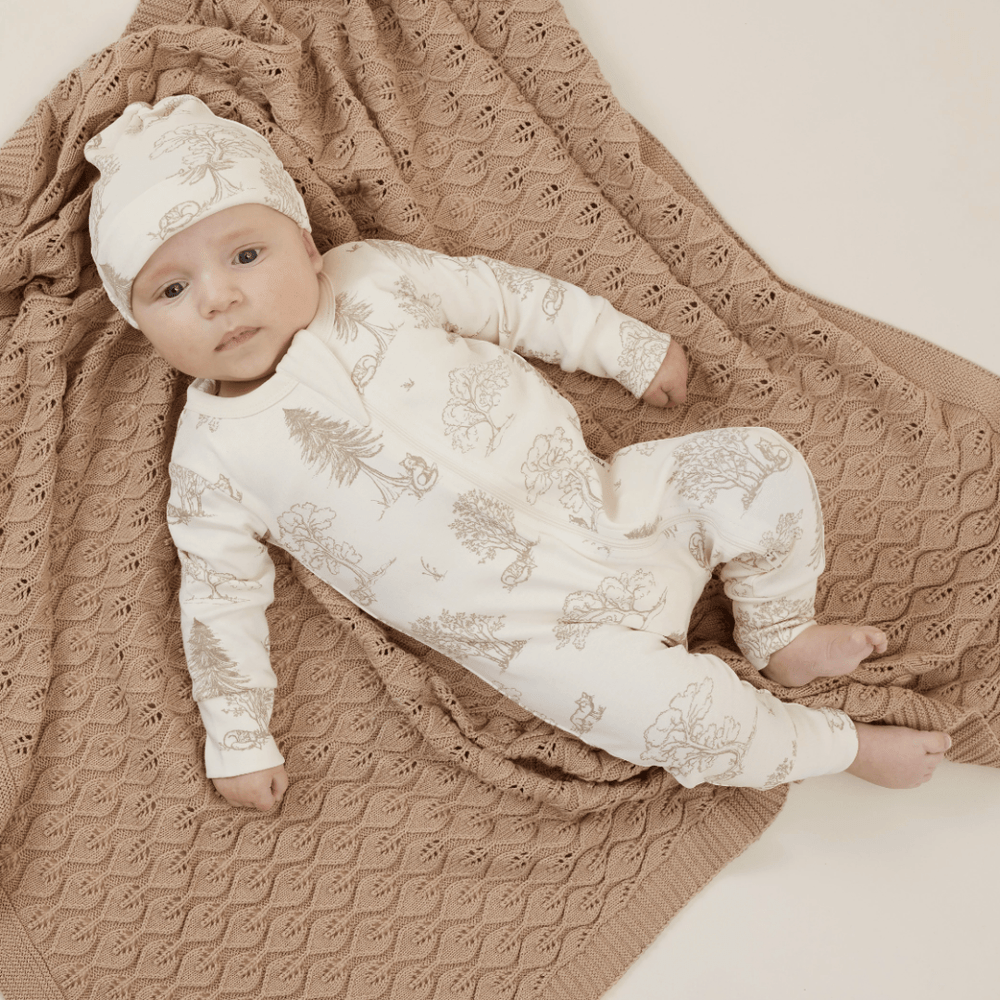 A baby wearing a white floral outfit and matching hat lies on an Aster & Oak Organic Leaf Knit Baby Blanket - LUCKY LAST, gazing upwards.