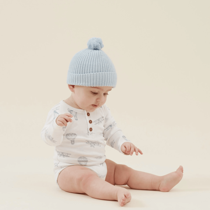 A baby in an Aster & Oak Organic Henley Long-Sleeved Onesie, LUCKY LASTS - FOX WOODS ONLY, paired with a blue knit hat, sits on a plain background.