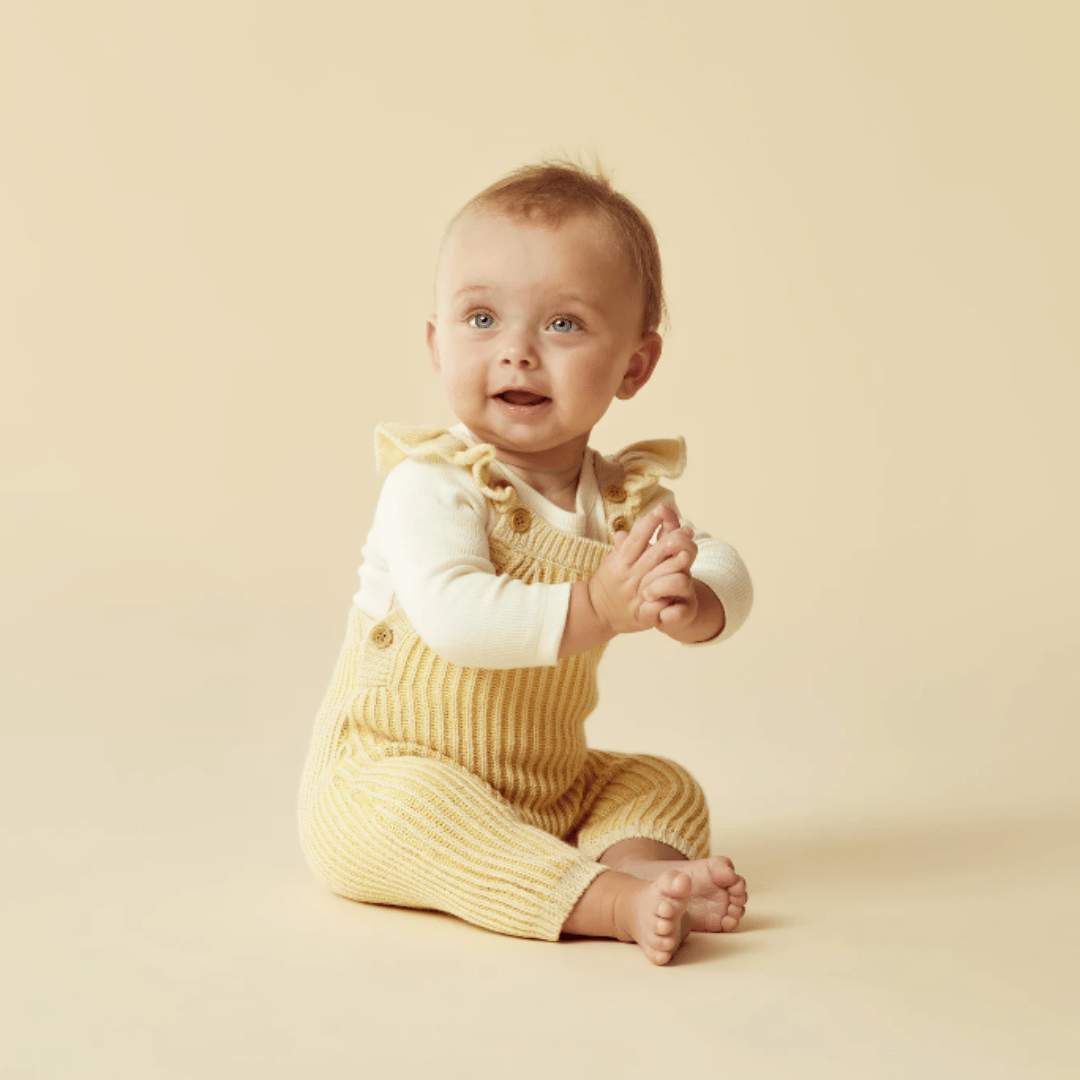 A baby sits on a light beige background, wearing a white long-sleeve shirt and Wilson & Frenchy's Knitted Ruffle Overalls - LUCKY LASTS - DIJON ONLY, with hands clasped together and looking upward.