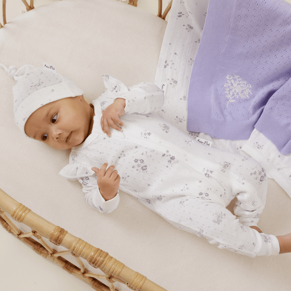 A baby in a white floral onesie and matching hat lies in a wicker bassinet next to an Aster & Oak Organic Ruffle Knit Baby Blanket by Aster & Oak.