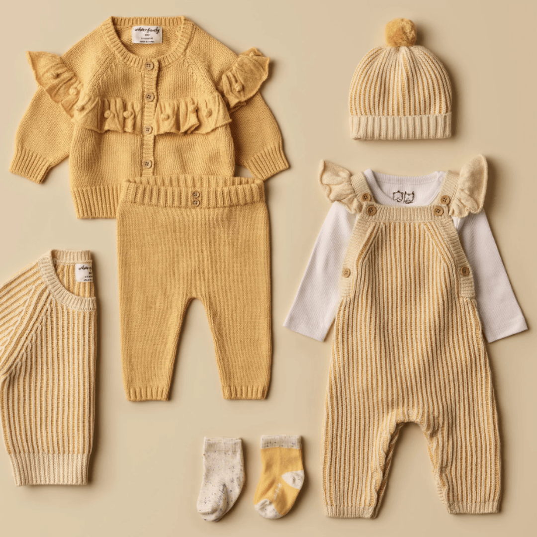 A collection of Wilson & Frenchy yellow knitted baby clothes, including cardigans, pants, a pom-pom hat, the adorable Wilson & Frenchy Knitted Ruffle Overalls - LUCKY LASTS - DIJON ONLY with a matching long-sleeve shirt and two pairs of socks, all arranged neatly on a beige background.