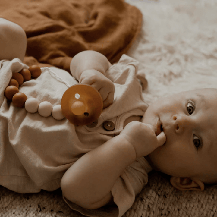 A baby lying on a soft surface holds the Funny Bunny Kids Silicone & Beechwood Dummy Clip, made of silicone beads, and has a finger in their mouth.