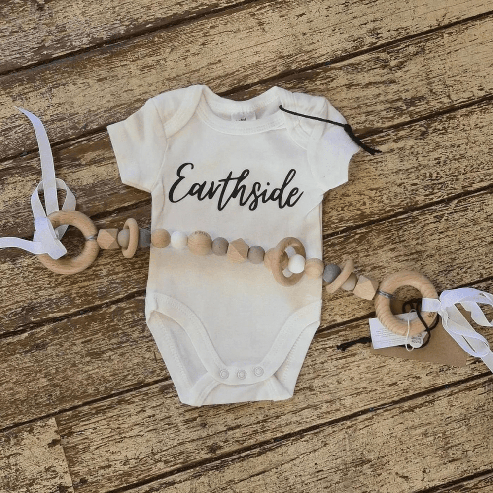 A charming baby onesie labeled "Earthside" rests on a wooden surface beside the delightful Funny Bunny Kids Silicone & Beechwood Pram Garland adorned with silicone beads and ribbons.