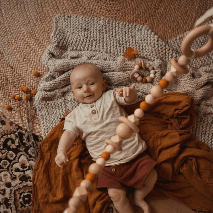 A baby rests on a textured blanket, surrounded by neutral-colored toys, including the adorable Funny Bunny Kids Silicone & Beechwood Pram Garland, while dried flowers lie nearby.