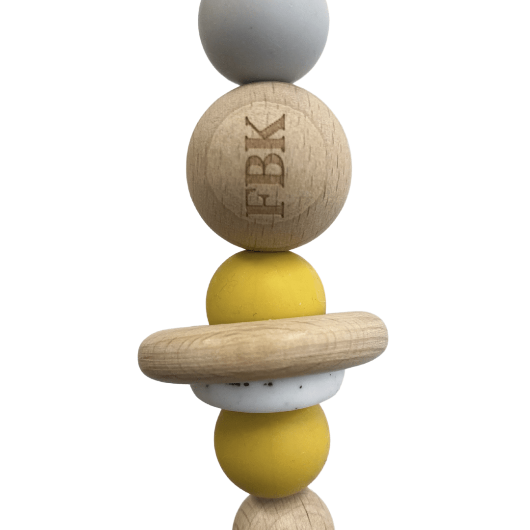 The Funny Bunny Kids Silicone & Beechwood Pram Garland features an arrangement of wooden and silicone beads in gray, yellow, and natural wood tones, resembling a charming baby toy. The top wooden bead is engraved with "FBK," making it perfect for adding a personal touch to your pram garland.