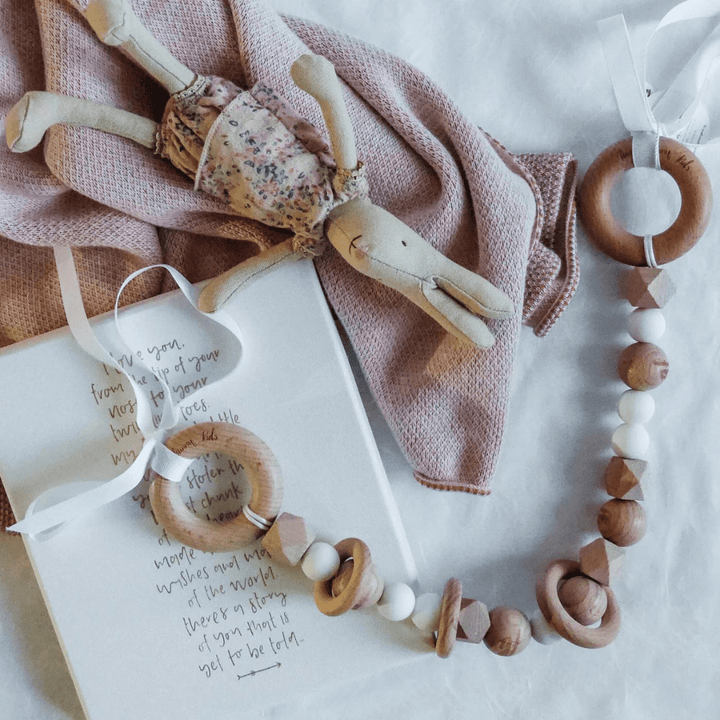 A stuffed rabbit toy lies on a pink blanket, accompanied by the Funny Bunny Kids Silicone & Beechwood Pram Garland and an open notebook with handwritten text.