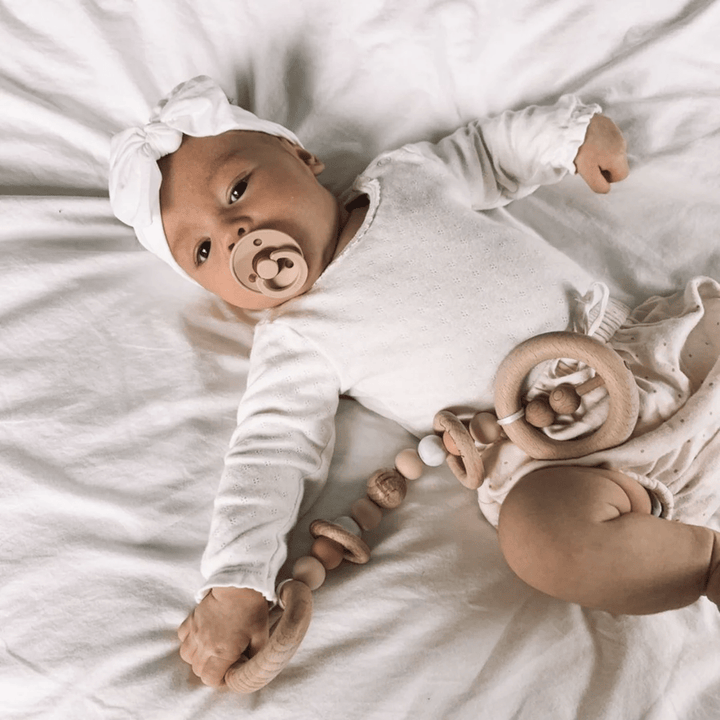 A baby lies on a white blanket, dressed in a white outfit and headband, clutching a wooden teething toy made of natural beechwood. With a pacifier in their mouth, the serene scene is complemented by the subtle elegance of Funny Bunny Kids Silicone & Beechwood Play Gym Toy nearby.