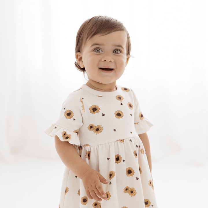 Smiling toddler girl with brown hair wearing an off-white dress with bright yellow sunflowers and ruffle sleeves