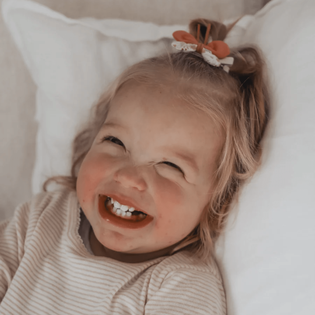 A young child with a big smile, rosy cheeks, and Over the Dandelions Butterfly Hair Clips lies on a pillow, looking up.