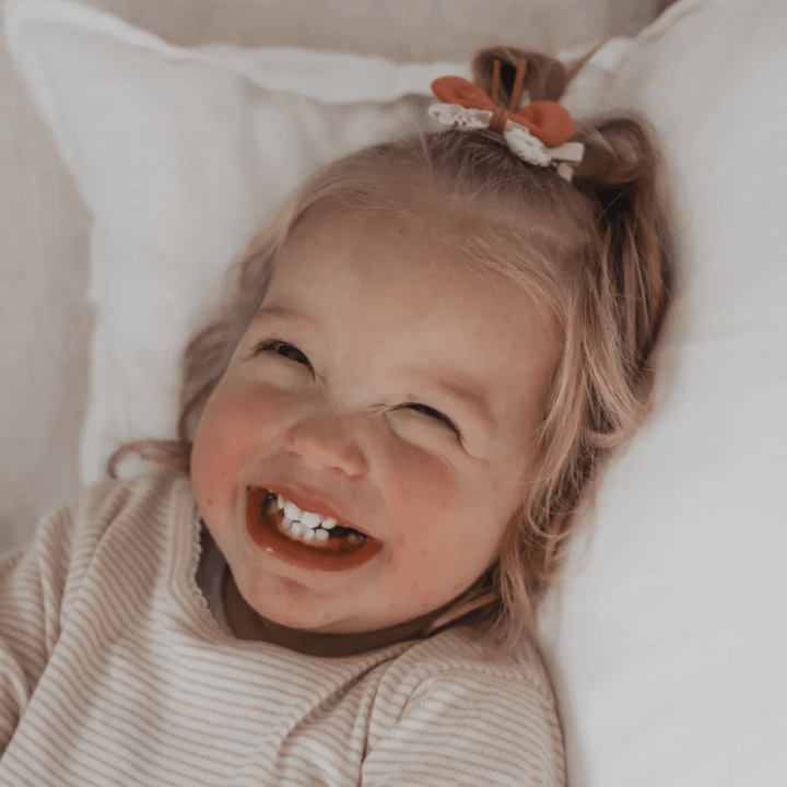 A young child with a big smile, rosy cheeks, and Over the Dandelions Butterfly Hair Clips lies on a pillow, looking up.