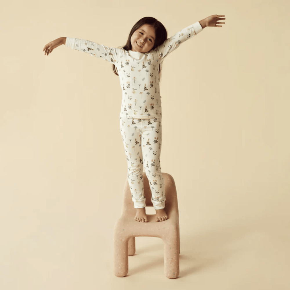 A child wearing Wilson & Frenchy Organic Long Sleeved Pyjamas stands on a beige chair with arms stretched out and a smile on their face.